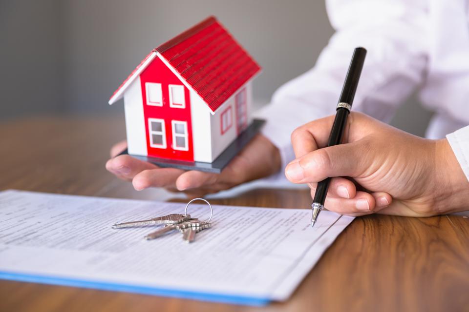 Hand signing document with a key on it and another hand giving a small house in return